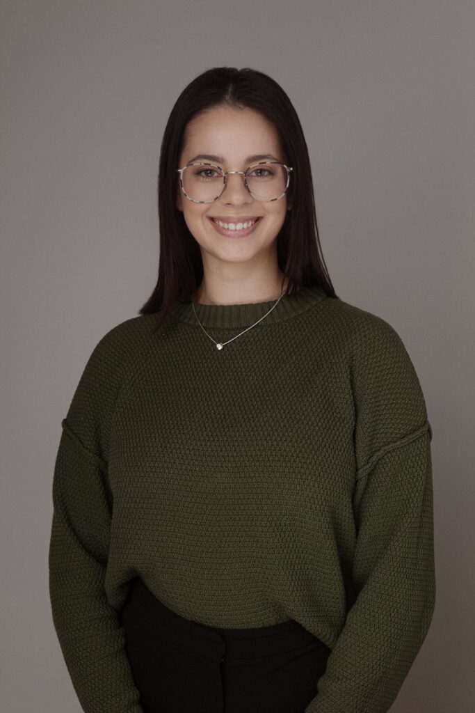 A smiling woman wearing a green sweater and glasses, standing against a neutral background. She is dressed professionally, representing Friday Health.