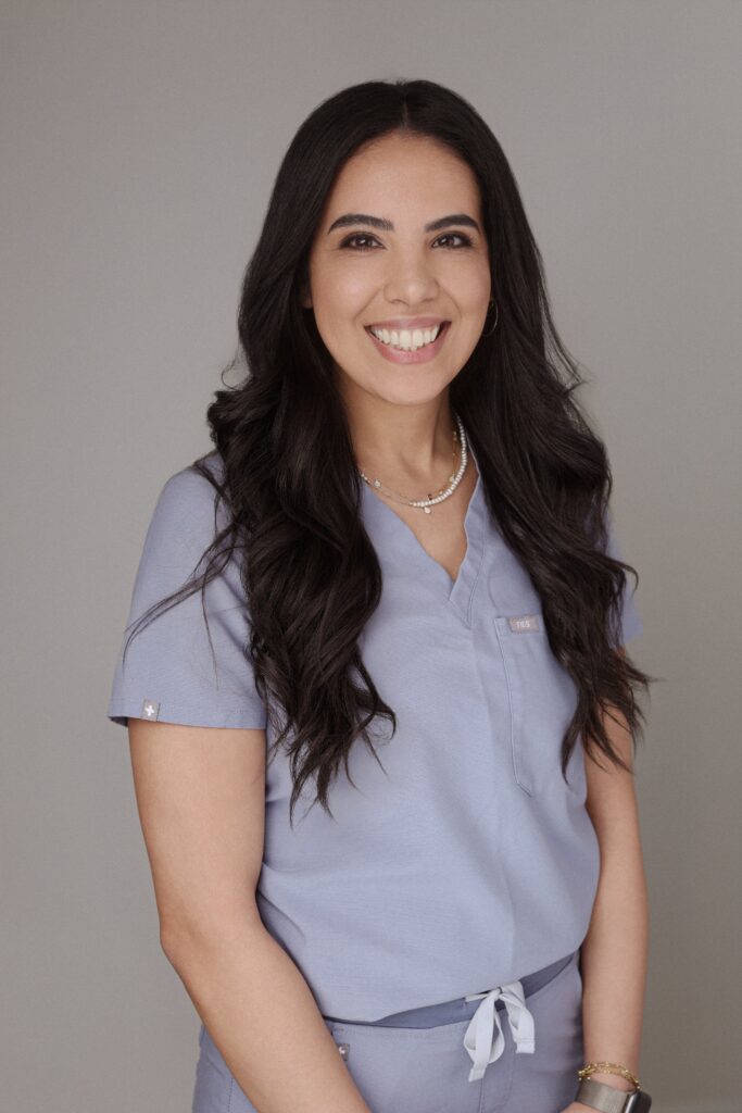 A smiling individual wearing light blue medical scrubs stands against a gray background, representing Friday Health. The person has long dark hair and accessorizes with a necklace, adding a professional yet friendly appearance.