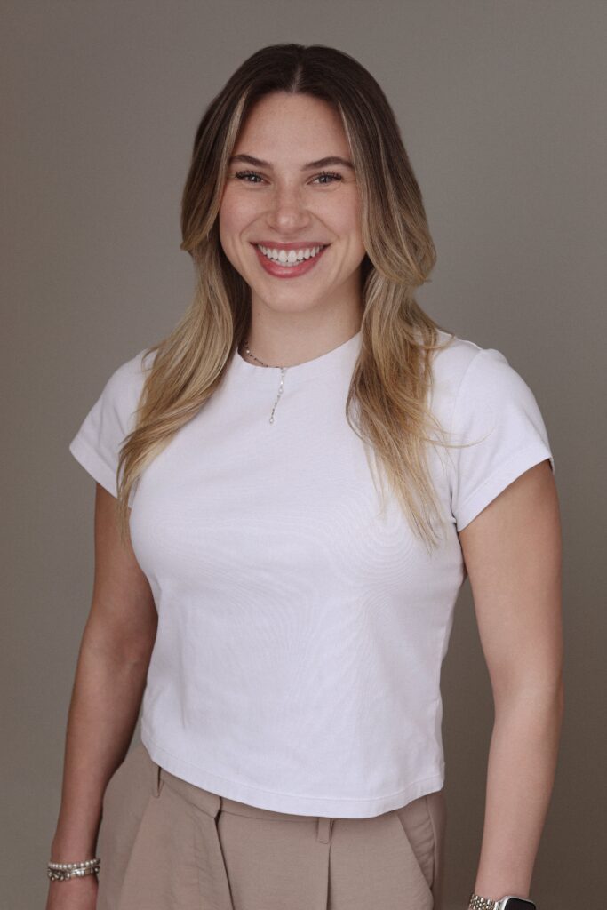 A smiling young woman wearing a white shirt and beige pants, standing against a neutral background. This image is associated with Friday Health.