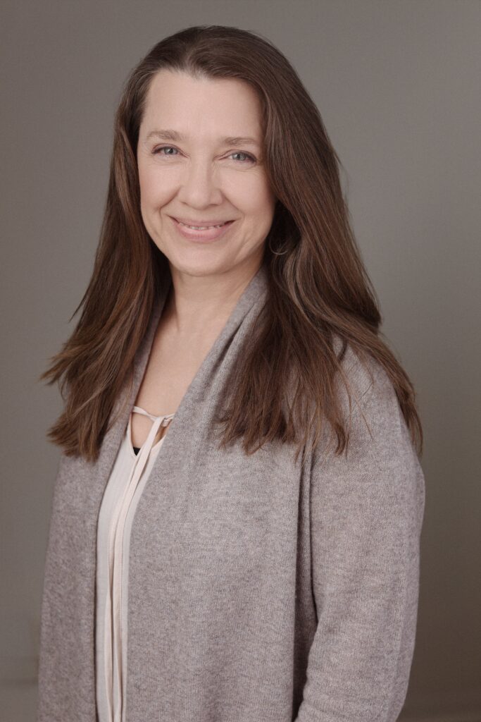 A professional-looking woman with long brown hair, wearing a light-colored top and a cozy cardigan, smiles warmly. The backdrop is a soft, neutral tone, complementing the theme of Friday Health.