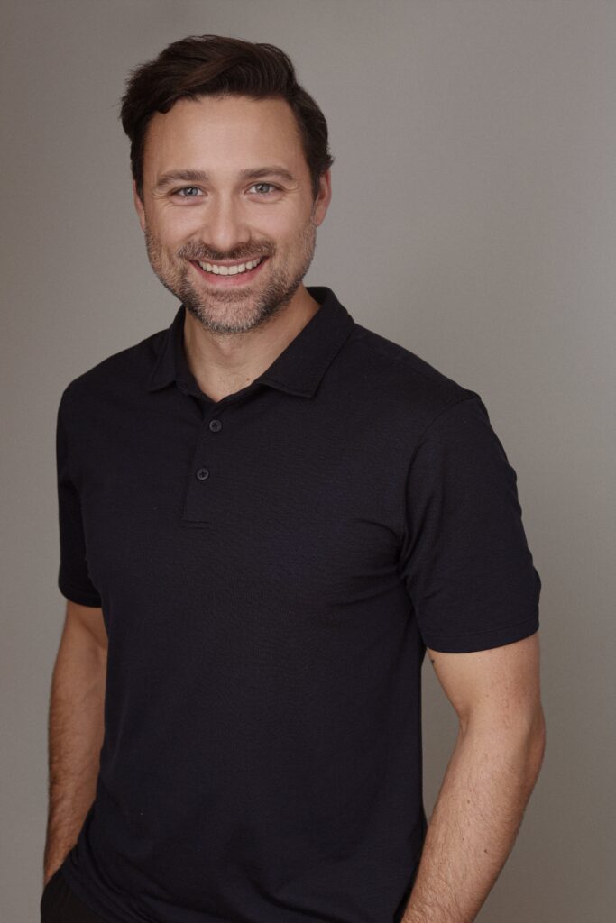 A smiling man with dark hair and a beard is wearing a black polo shirt, standing against a neutral background. His expression conveys friendliness and approachability, representing the values of Friday Health.