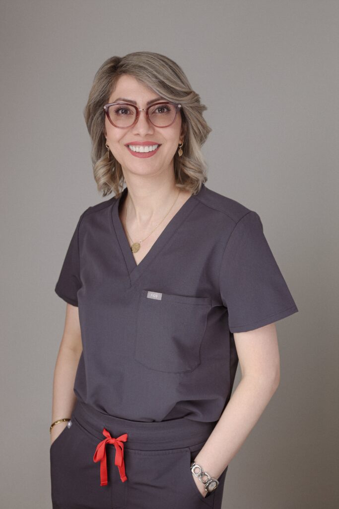 A smiling professional wearing dark scrubs with a red tie at the waist and glasses, posed against a neutral gray background, representing Friday Health.