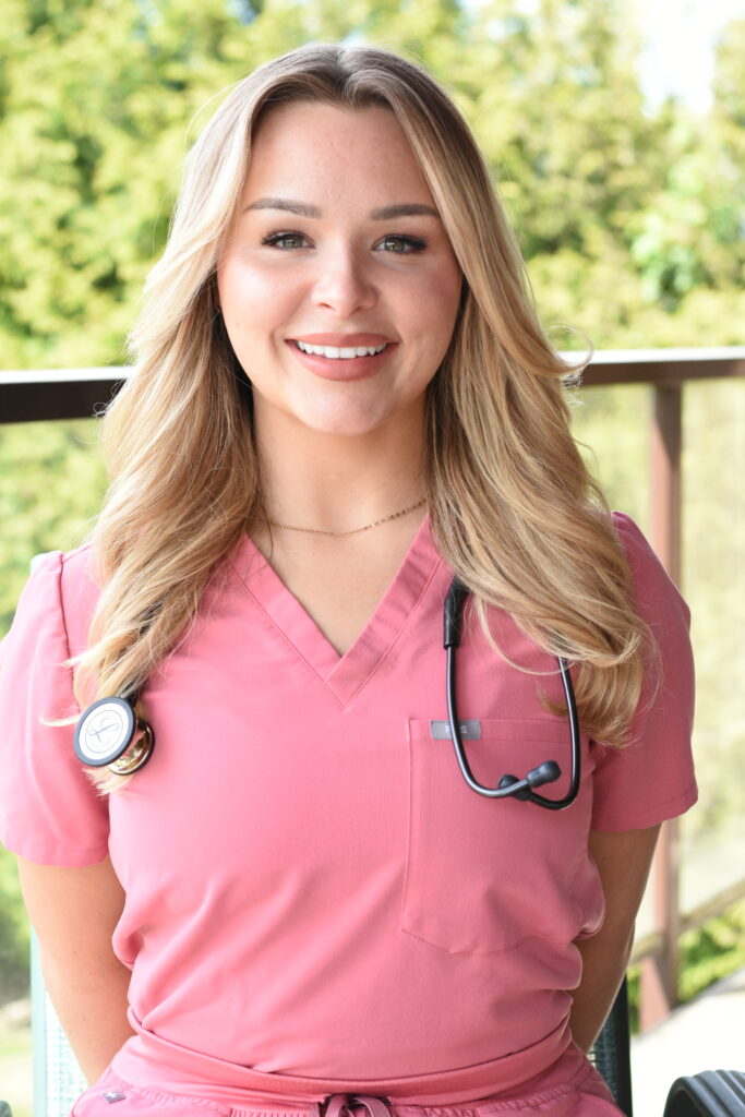 A healthcare professional in pink scrubs smiles warmly, wearing a stethoscope around her neck, symbolizing the compassionate care associated with Friday Health. The background features a lush green landscape, adding a refreshing touch to the scene.