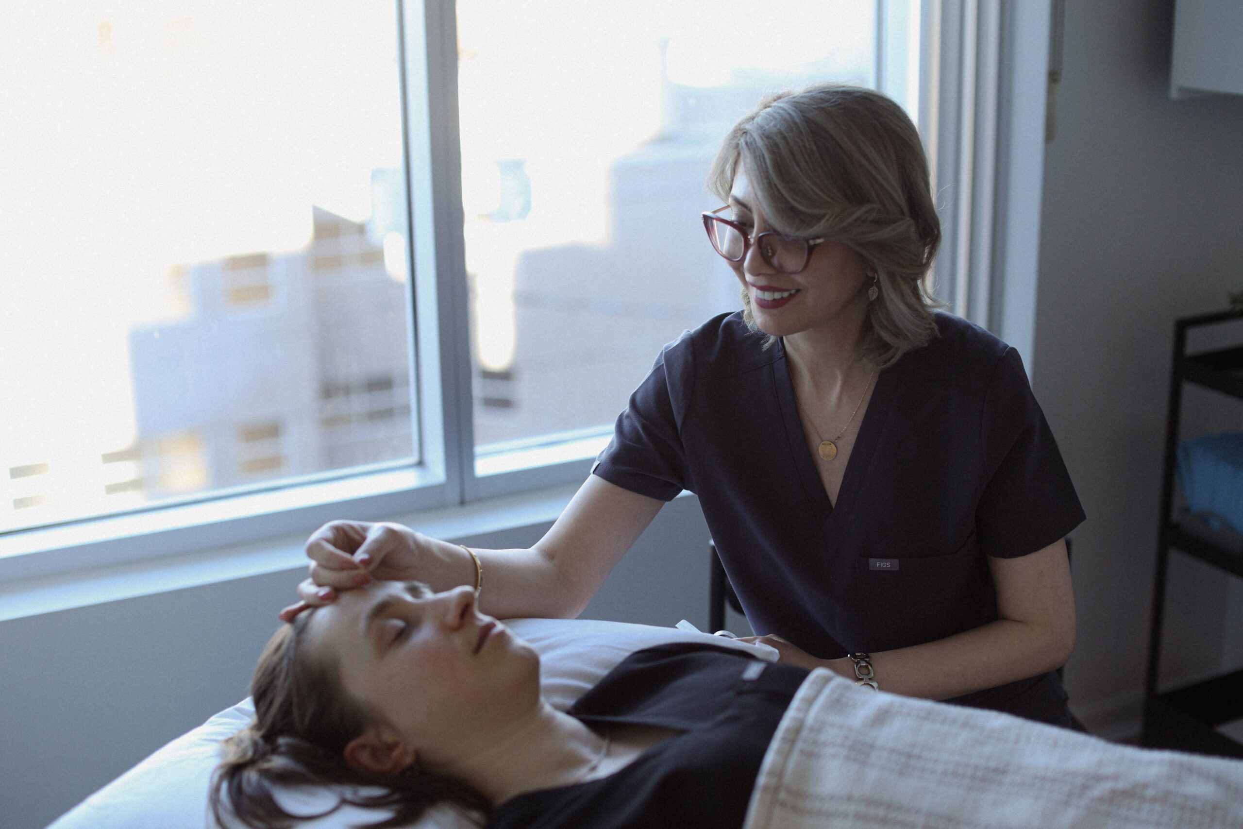 A healthcare professional at Friday Health is gently applying a treatment to a patient's forehead while the patient relaxes on a treatment table, with a bright window and city view in the background.