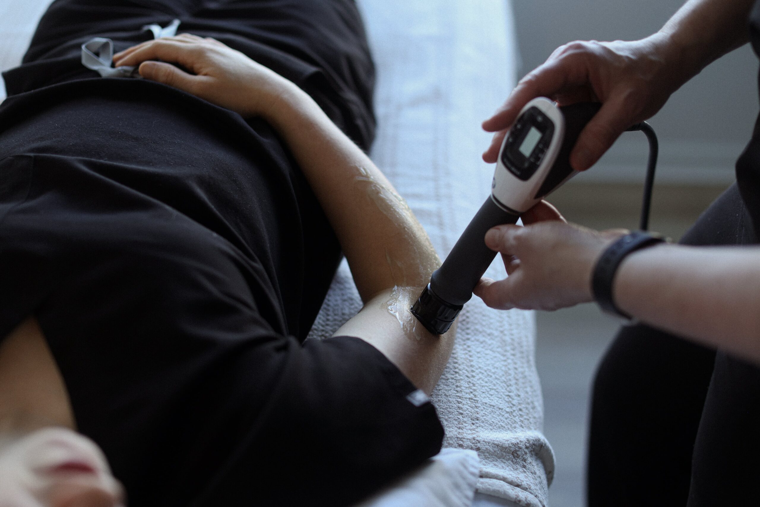 A person is lying on a treatment table with their arm exposed. Another person is applying a device to the arm, which appears to be covered with a gel. This scene is taking place in a clinic related to Friday Health services.