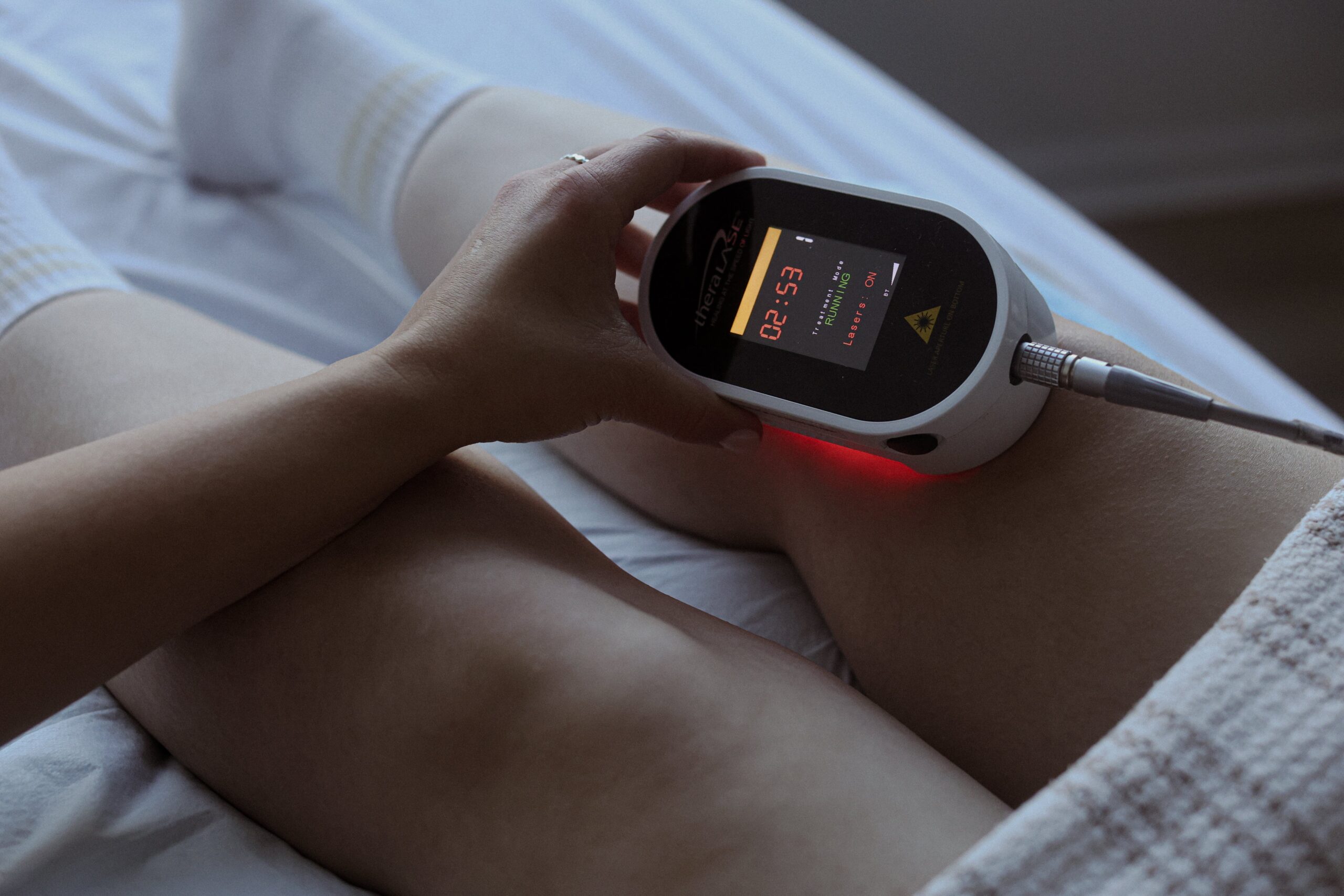 A person uses a handheld device for therapeutic treatment on their leg while resting on a bed, with a focus on health and wellness, highlighting the importance of Friday Health.