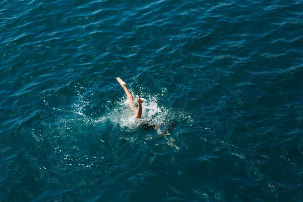 A person is diving into a bright blue ocean, creating splashes of water, with their legs visible in the air, symbolizing the spirit of adventure and wellness promoted by Friday Health.