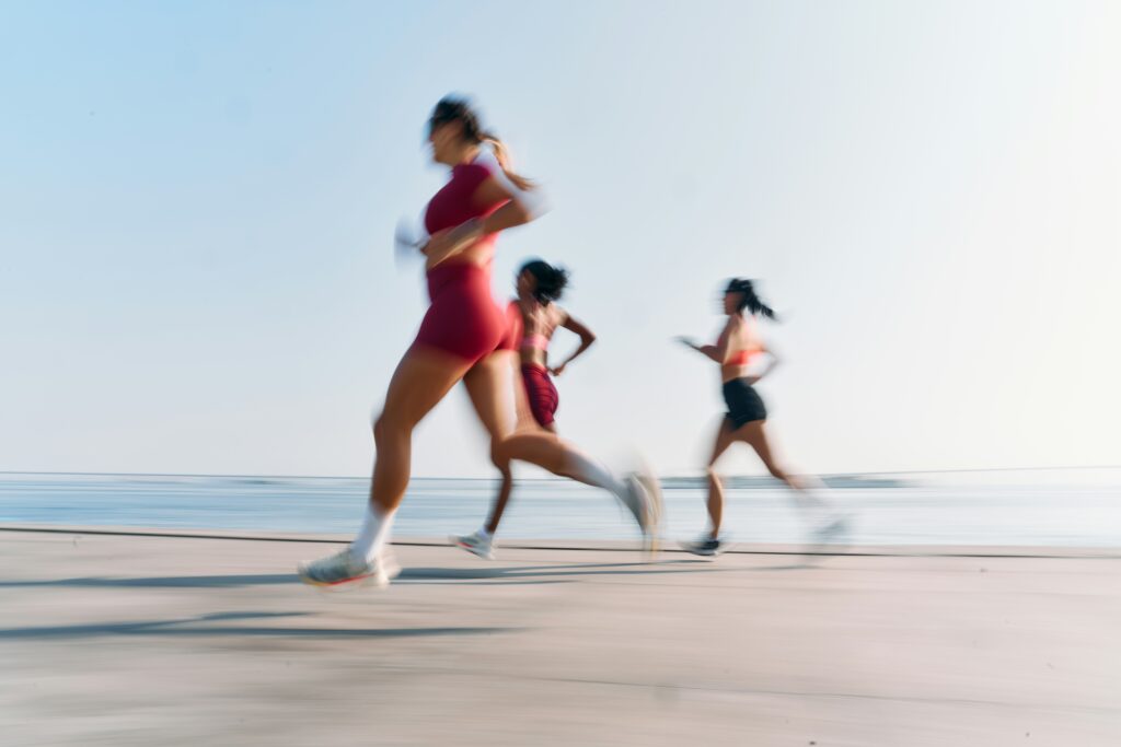 Three women are running along a beachside path, showcasing an active lifestyle associated with Friday Health. The image conveys a sense of motion and energy against a bright sky and ocean backdrop.