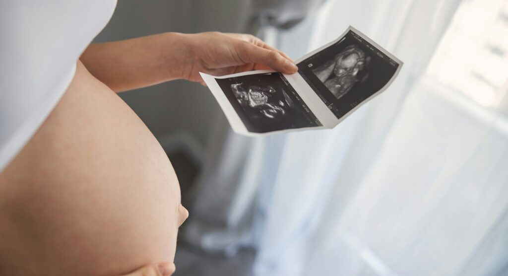 A pregnant person holds ultrasound images while looking at their baby bump, emphasizing the connection to Friday Health.