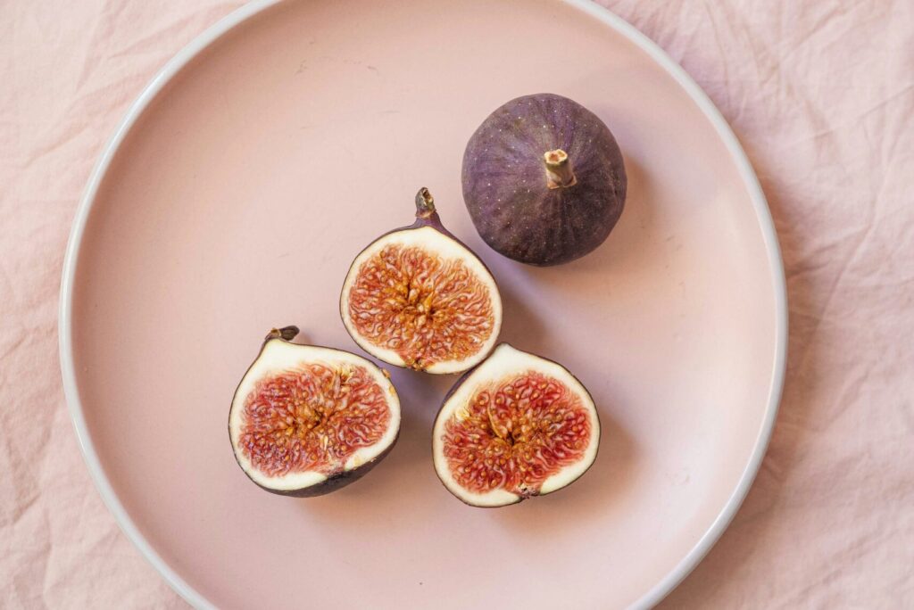 Alt text: A pink plate featuring two halved figs with a single whole fig beside them, set against a soft pink fabric background, emphasizing a fresh, health-conscious vibe associated with Friday Health.