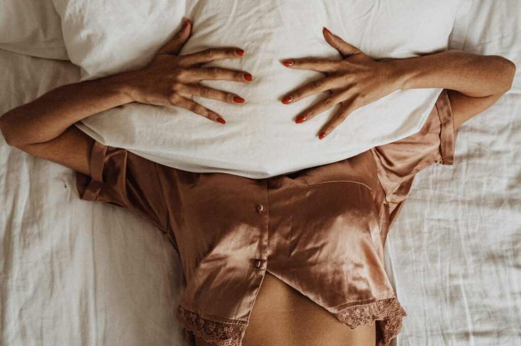 A person wearing a satin outfit is lying on a bed, holding a pillow over their face, surrounded by white bedding. The scene evokes a sense of relaxation and comfort, tying into the theme of Friday Health and self-care.