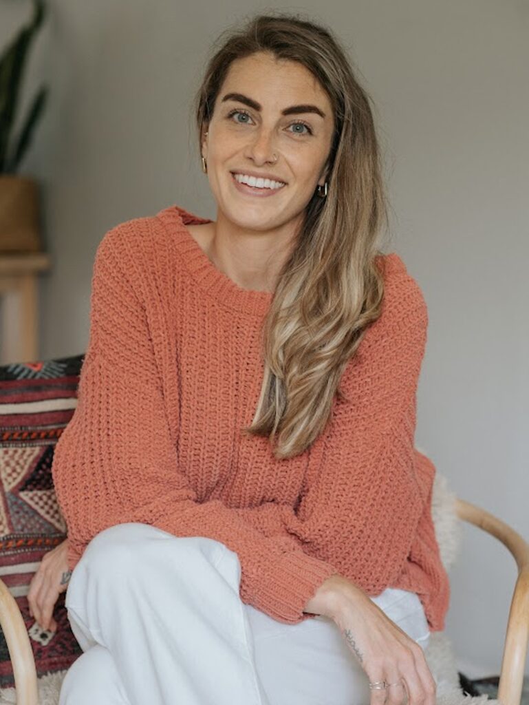 A woman sits in a cozy chair, smiling warmly at the camera while wearing a soft orange sweater and white pants. In the background, there is a plant and a patterned blanket. The image conveys a sense of comfort and health, embodying the essence of Friday Health.