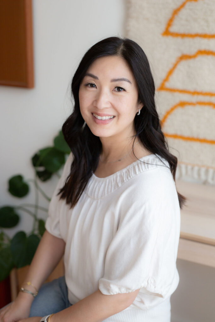 A woman with dark hair is smiling while sitting in a bright room decorated with plants and a textile wall hanging. The image represents a positive atmosphere associated with Friday Health.