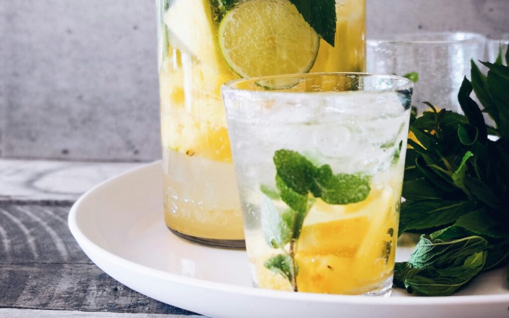 A refreshing drink made with mint, lime, and pineapple, served in a glass alongside a jar of the same beverage, placed on a white plate. This setup emphasizes the theme of hydration and wellness, in line with Friday Health.