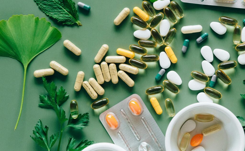 Flat lay of assorted pills and herbs on green surface, symbolizing health and wellness.