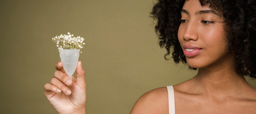 Happy young African American female model with curly hair in bra smiling and showing menstrual cup with gentle flowers against beige background
