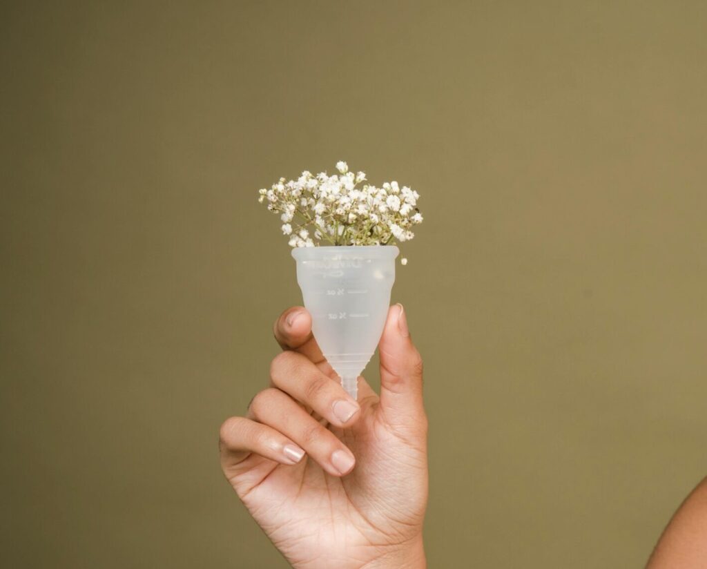 Happy young African American female model with curly hair in bra smiling and showing menstrual cup with gentle flowers against beige background