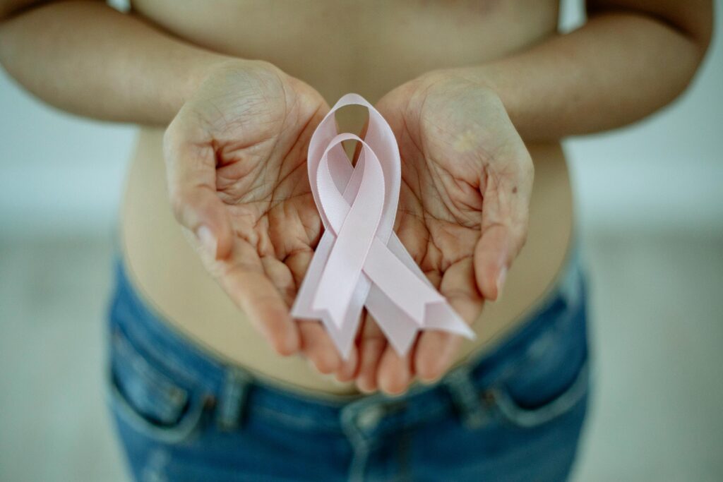 High angle of crop unrecognizable topless lady showing pink ribbon in arms as symbol of breast cancer awareness while standing in bright room